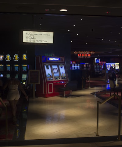 A dimly lit casino with several rows of slot machines, some displaying bright graphics and lights. There are a few people scattered throughout the area, engaging with the machines. The floor is reflective and polished, adding to the ambiance, while the ceiling is dotted with numerous small lights.