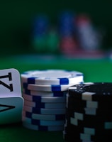 A casino scene featuring two playing cards, the ace and king of clubs, partially concealed by a player's hand. Nearby, there are stacks of multicolored poker chips on a green felt table, with blurred colorful background elements suggesting other chip stacks or cards.